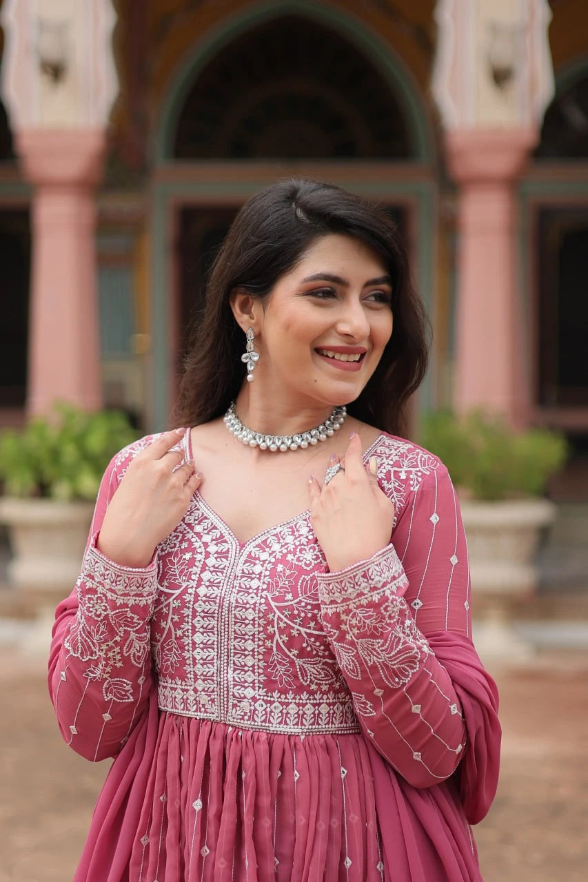 Woman in a pink traditional outfit with intricate designs, standing in front of an architectural background.
